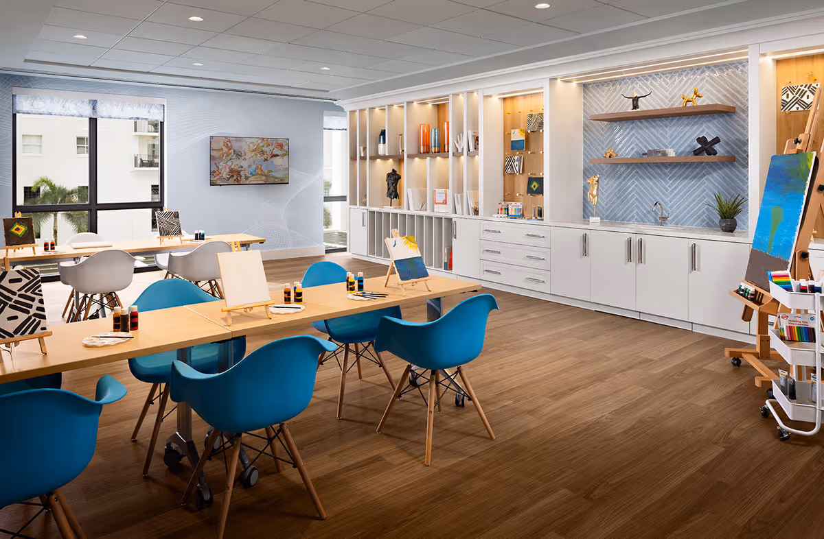 A bright and spacious art room with wooden floors, light blue and white walls, and large windows letting in natural light. The room features several tables with blue and white chairs, easels with blank canvases and some with paintings, and art supplies like paint bottles and palettes. Built-in white cabinets and shelves along one wall display decorative items and additional art materials.