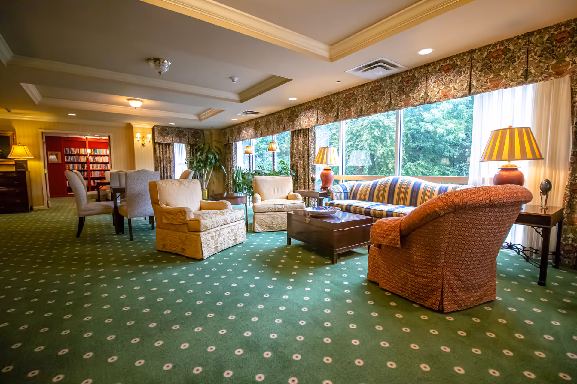 A cozy senior living community lounge area with patterned green carpet, a striped sofa, several armchairs, wooden coffee tables, and lamps. Large windows with floral curtains allow natural light and offer a view of green trees outside. In the background, there is a dining area with chairs and a bookshelf against a red wall.