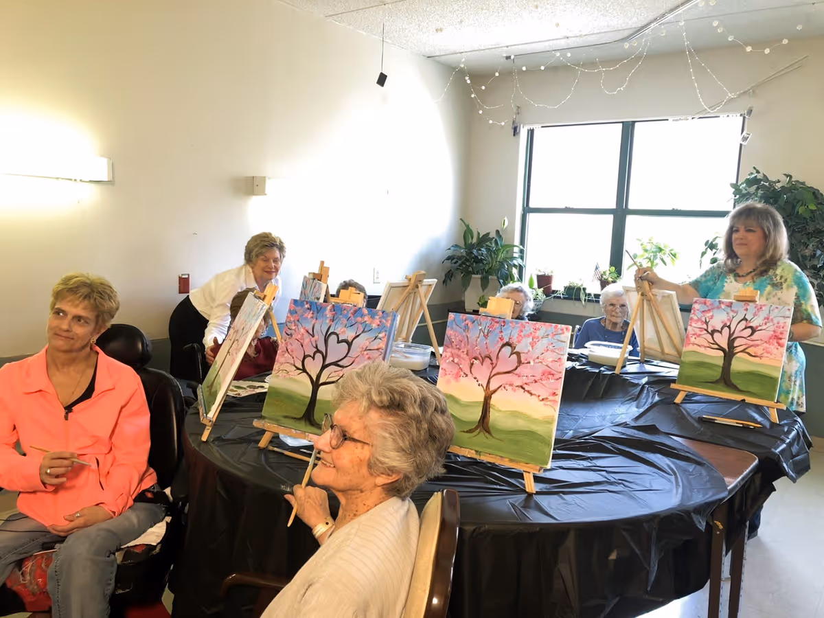 A group of elderly women and a younger woman participating in a painting activity around a large round table covered with a black plastic tablecloth. Each person has an easel with a canvas depicting a tree with pink blossoms and a heart shape in the branches. The room has a window with plants on the windowsill and string lights hanging from the ceiling.