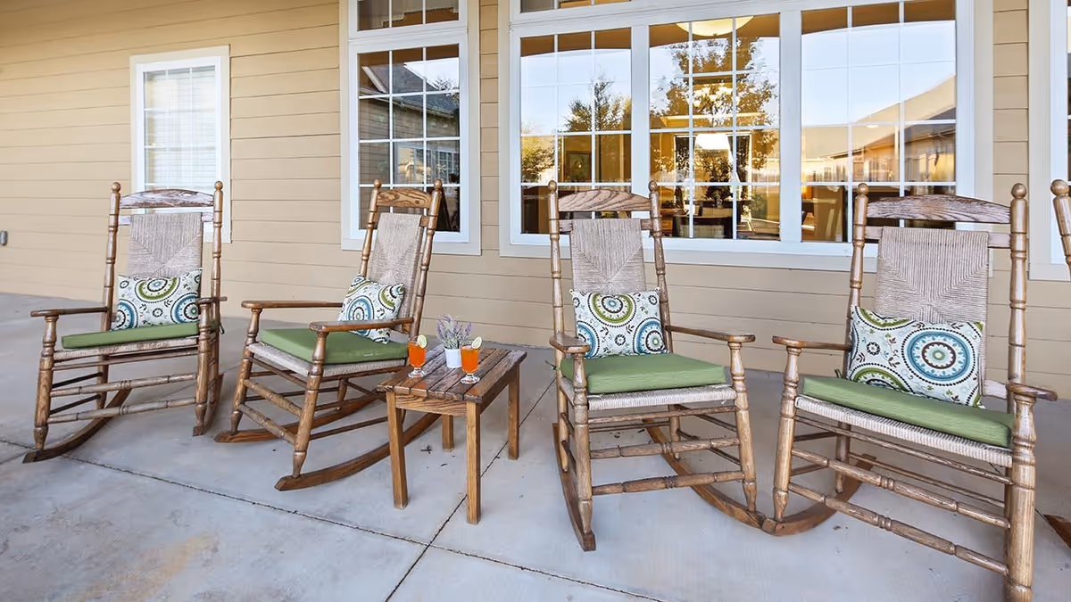 Four wooden rocking chairs with green seat cushions and patterned pillows are arranged on a concrete patio in front of a beige building with white-framed windows. A small wooden table between the chairs holds two orange drinks and a small decorative item.