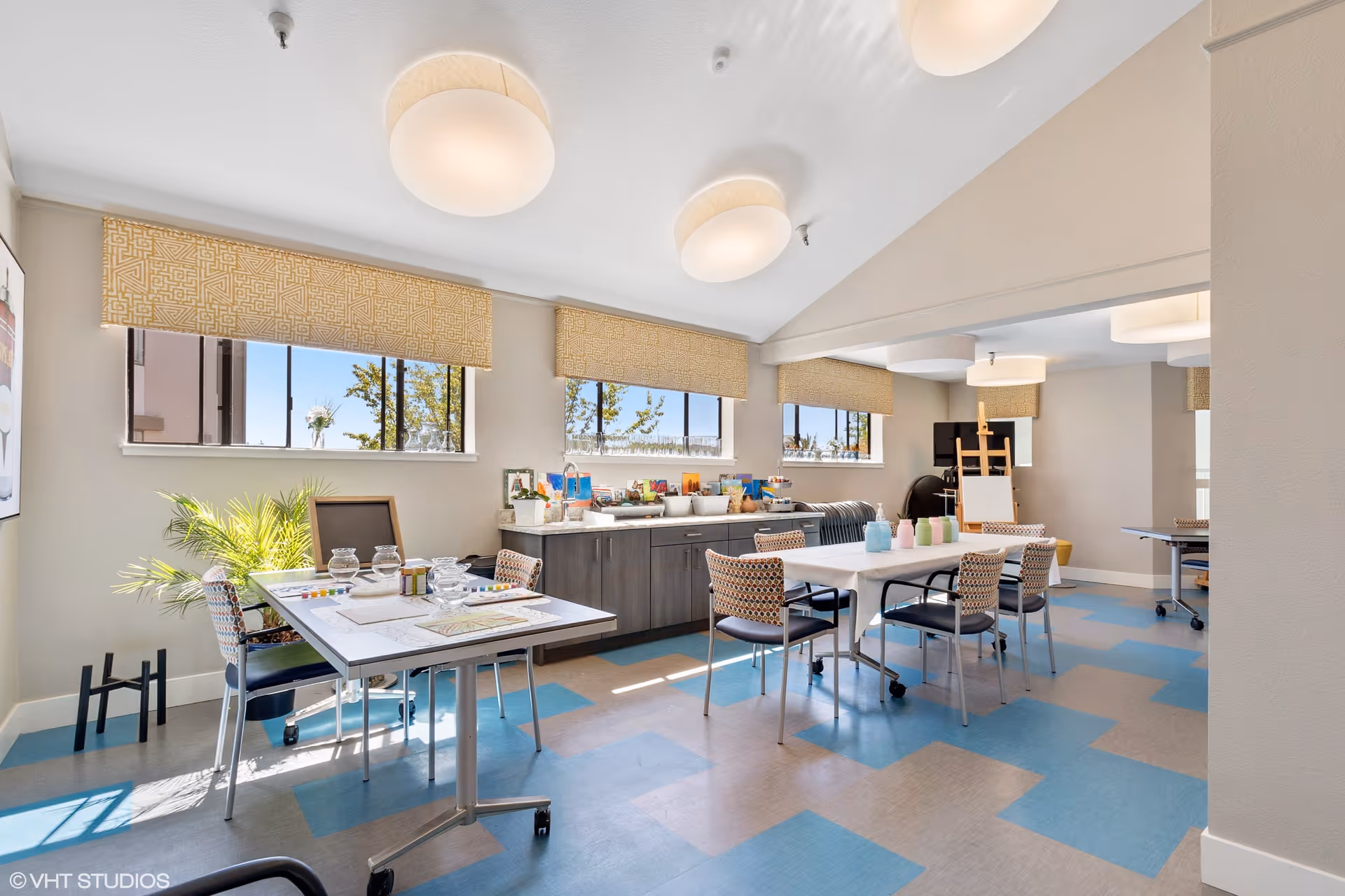 A bright and spacious activity room with large windows letting in natural light. The room features tables and chairs arranged for group activities, with art supplies and craft materials on the tables and a cabinet against the wall. The floor has a patterned design in blue and beige, and the ceiling has round light fixtures.
