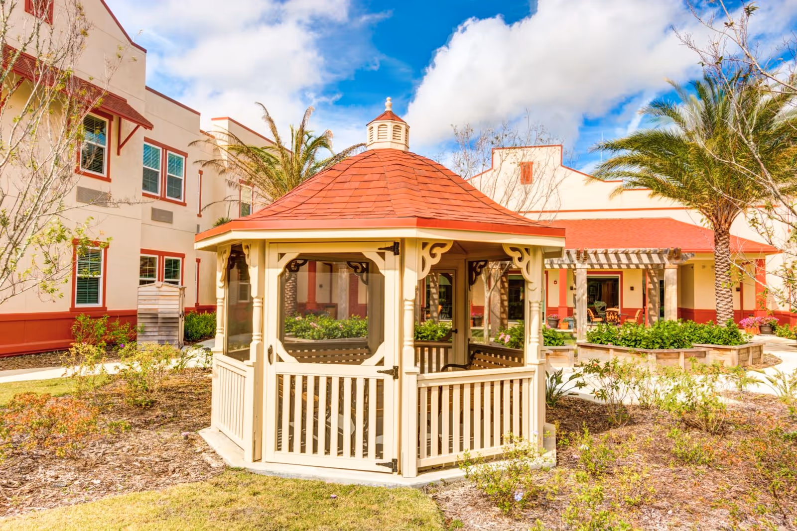 A beige wooden gazebo with a red shingled roof in a landscaped courtyard outside a senior living building.