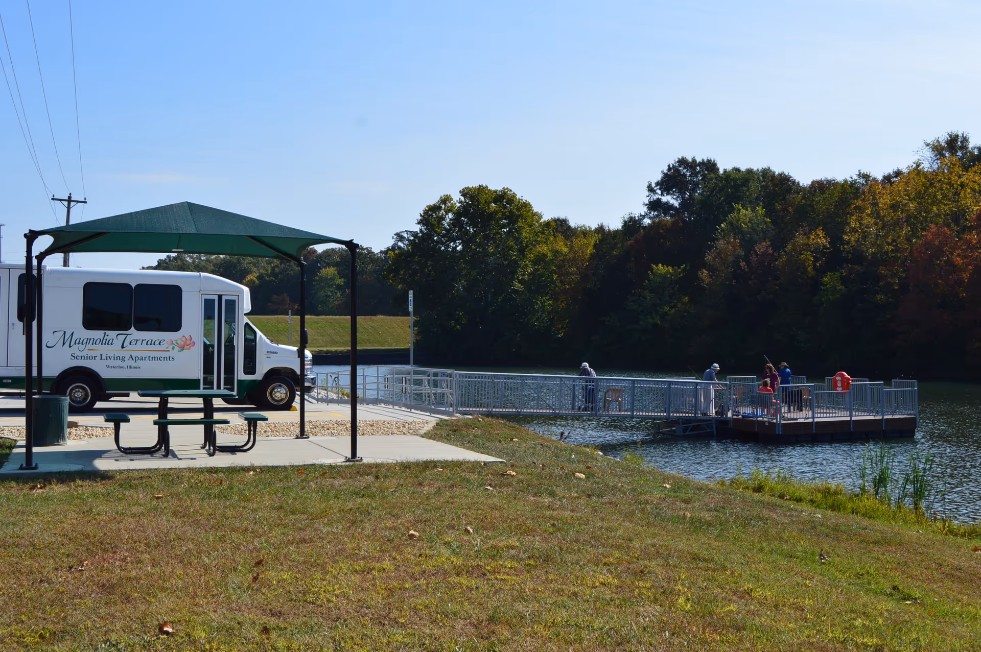 Outdoor scene at Magnolia Terrace Senior Living Apartments featuring a white shuttle bus with the facility's name parked near a green shaded picnic table. In the background, several people are standing on a metal dock extending into a calm lake surrounded by trees with autumn foliage.