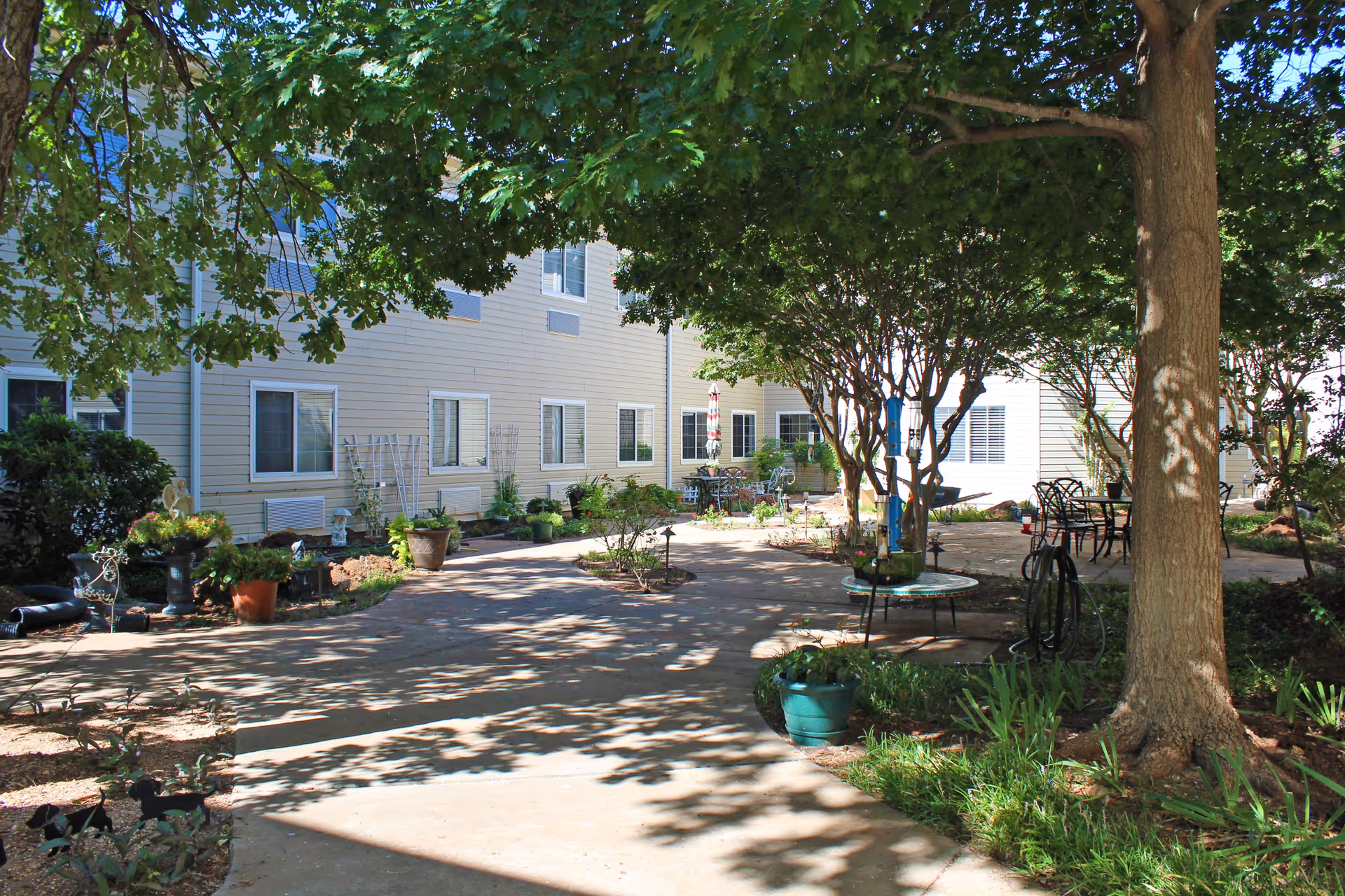 A shaded outdoor courtyard area at a senior living facility with a concrete pathway, potted plants, trees, and outdoor seating including tables and chairs. The building exterior is visible with multiple windows and light-colored siding.