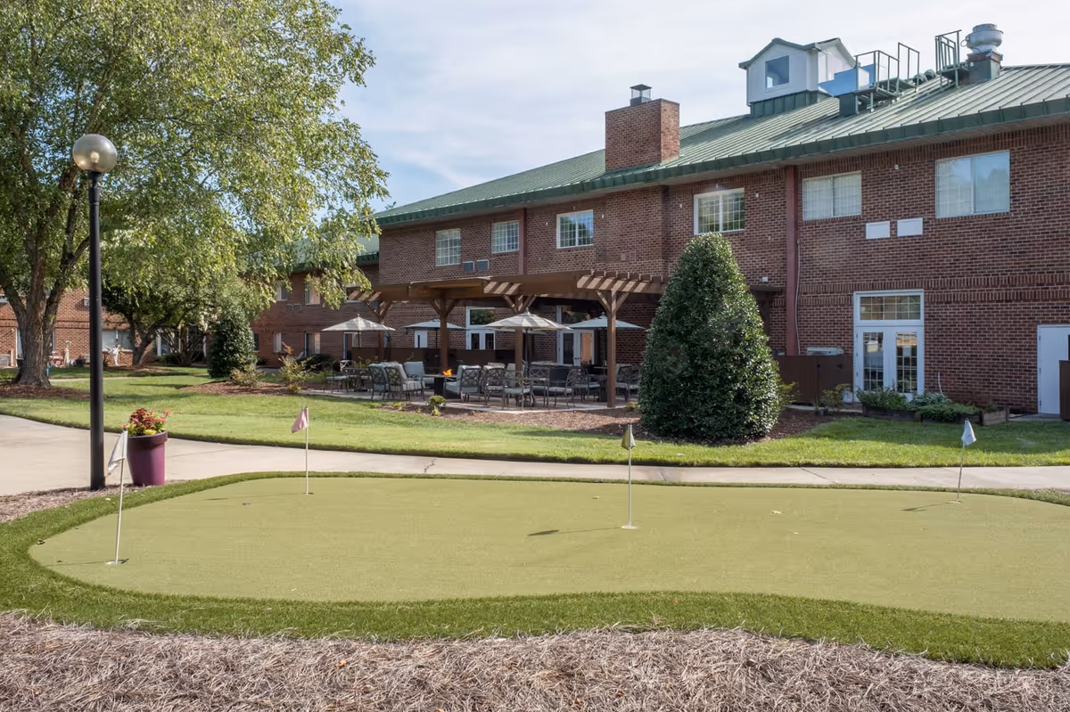 Outdoor view of a senior living facility with a putting green in the foreground, a paved walkway, and a brick building with a green roof and outdoor seating area under wooden pergolas. Trees and shrubs surround the area under a partly cloudy sky.