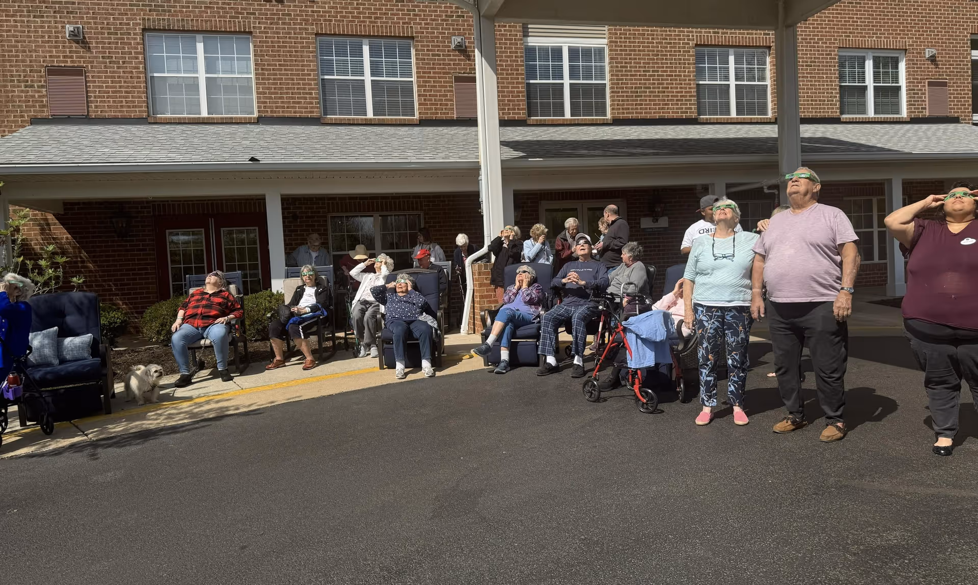 A group of elderly people sitting and standing outside a brick building under a covered area, all looking up at the sky wearing protective glasses, likely watching a solar eclipse. Some are seated in chairs or wheelchairs, and a small white dog is visible on the left side.