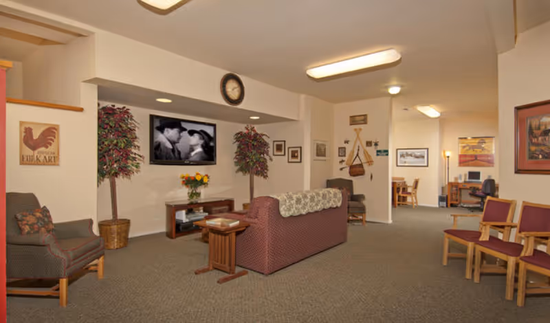 A cozy living room area in a senior living facility with a maroon couch, armchairs, and wooden chairs arranged around a TV mounted on the wall showing a black and white movie. The room has beige walls decorated with framed pictures, two potted plants, a clock above the TV, and a small table with a vase of flowers. In the background, there is a desk with a computer and additional seating.
