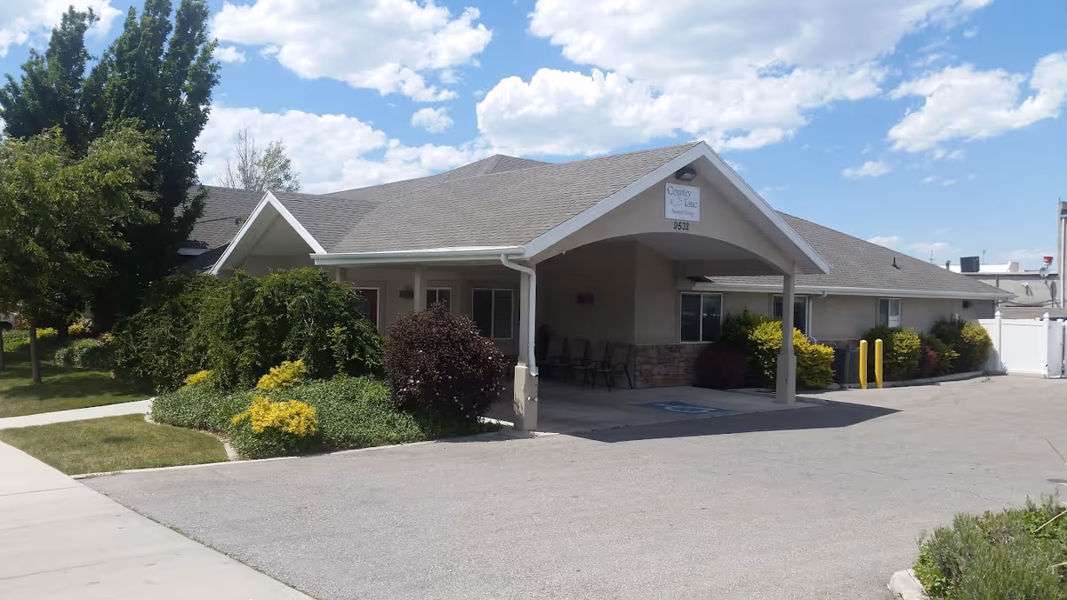 Exterior view of a single-story building with a covered entrance and a sign that reads 'Country Lane Assisted Living 9532'. The building is surrounded by well-maintained bushes, trees, and a paved driveway under a partly cloudy sky.