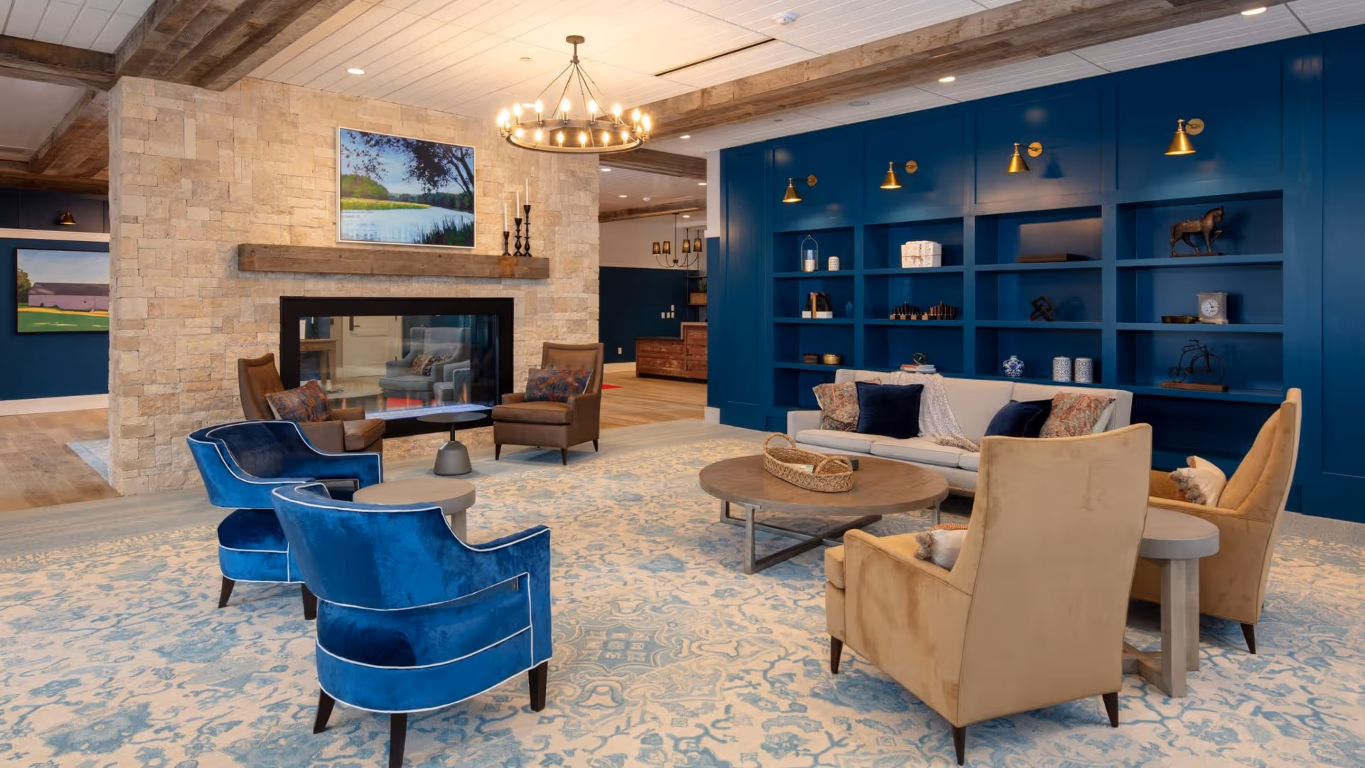 A cozy living room with a large patterned rug, two blue velvet armchairs, two beige armchairs, a white sofa with pillows, and a round wooden coffee table. The room features a stone fireplace with a painting above it, a blue built-in bookshelf with decorative items, wooden ceiling beams, and warm lighting fixtures.