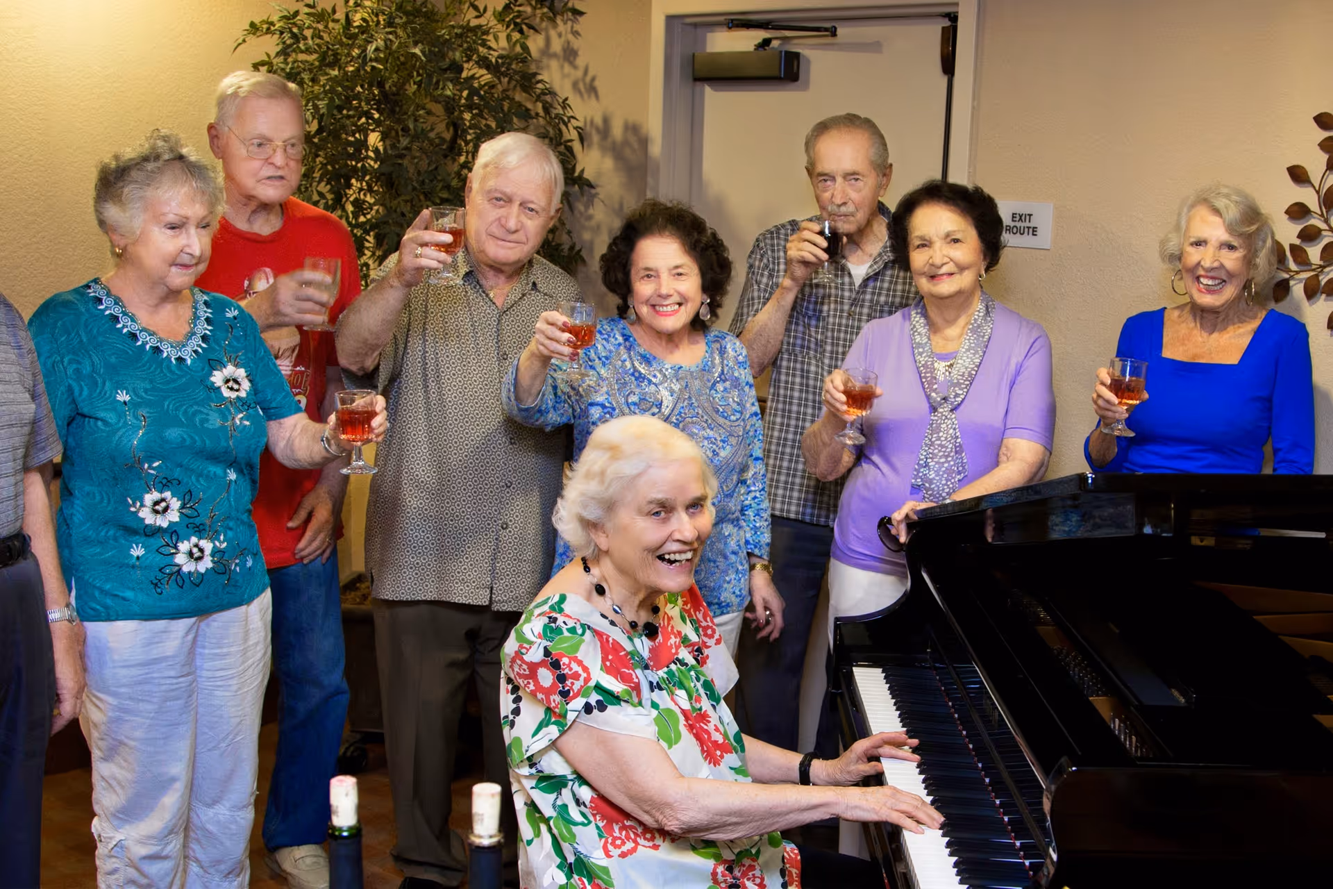 Several elderly people gathered around a grand piano in a communal room, smiling and raising glasses.