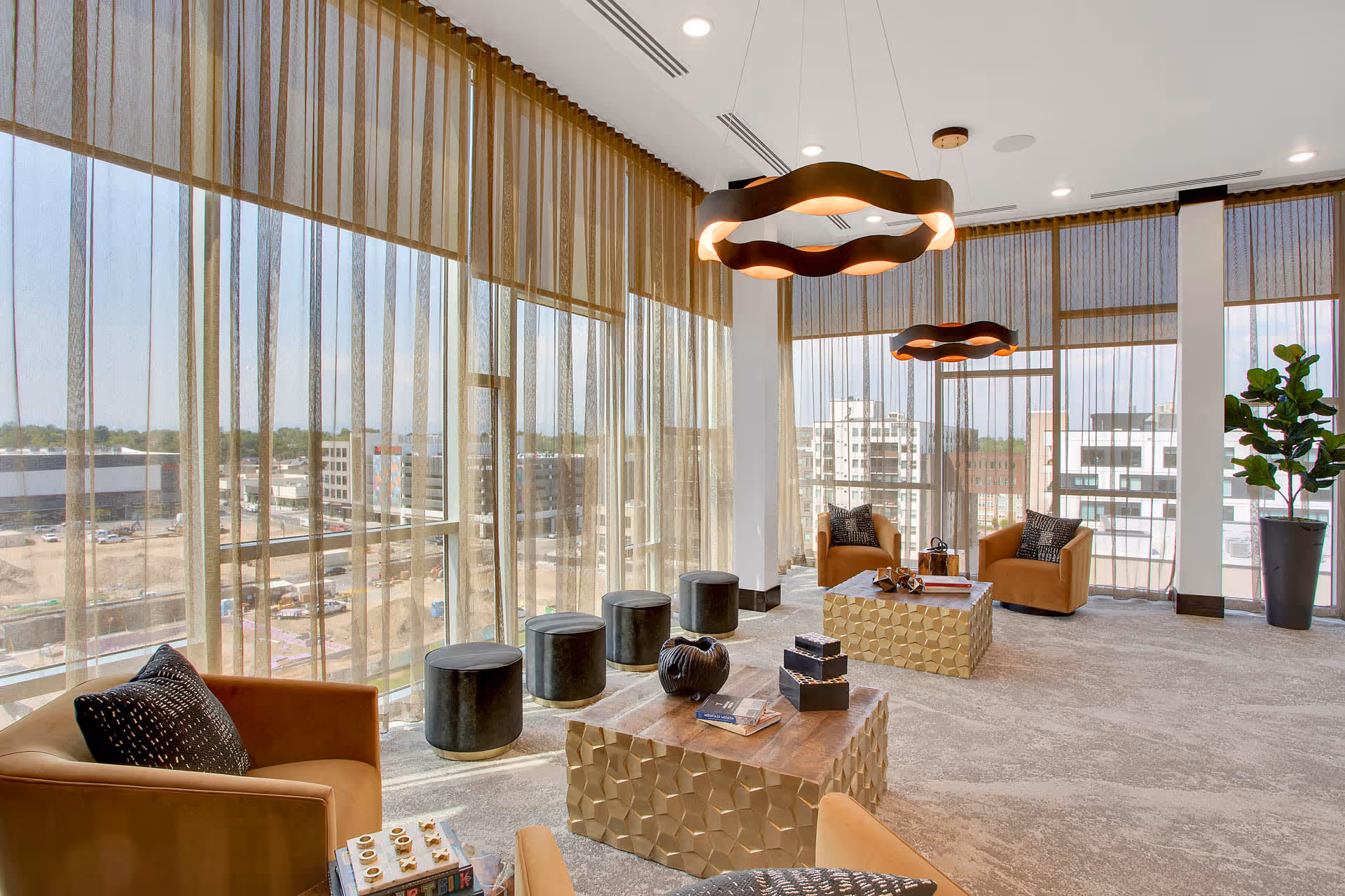 A modern lounge area with large floor-to-ceiling windows covered by sheer brown curtains. The room features tan armchairs with black and white patterned pillows, two gold geometric coffee tables with decorative items and books, and a row of black cylindrical stools near the windows. Two unique circular pendant lights hang from the ceiling, and a tall potted plant is placed in the corner. The view outside shows a cityscape with buildings and a clear sky.