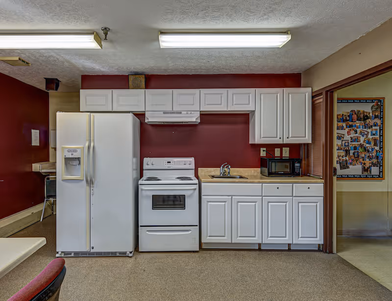 A kitchen area with white cabinets, a white refrigerator with an ice and water dispenser, a white electric stove with an overhead range hood, a countertop with a sink, and a microwave. The walls are painted dark red and beige, and there is a bulletin board with photos visible through an open doorway on the right.
