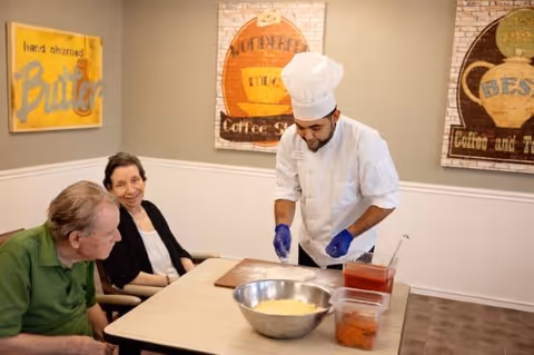 A chef in white prepares food at a table for two seated elderly residents in a dining room decorated with coffee-themed wall art.
