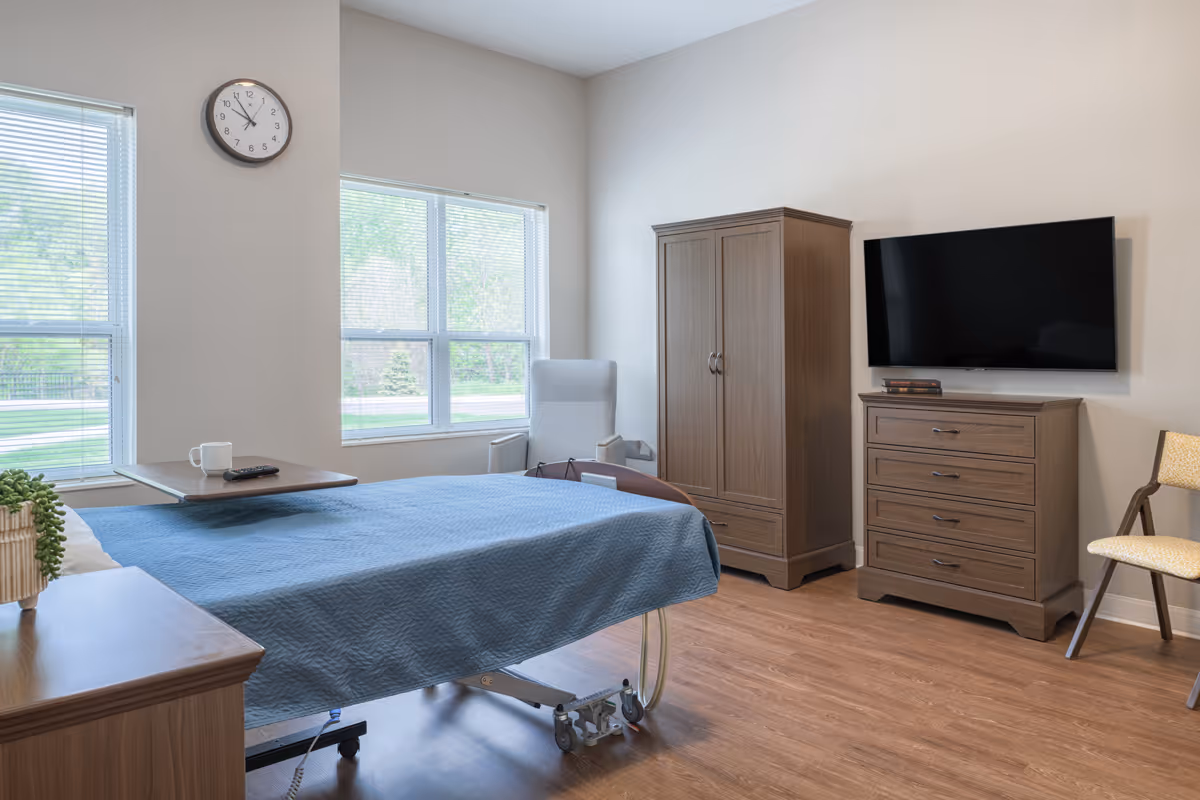 A senior living facility bedroom with a hospital-style bed covered in a blue quilt, a wooden nightstand with a plant, a wall clock, two large windows with blinds, a gray armchair, a wooden wardrobe, a wooden dresser with a flat-screen TV mounted above it, and a beige chair on a wooden floor.