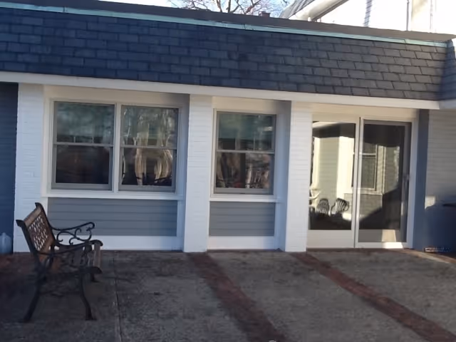 Exterior view of a building with two windows and a sliding glass door. There is a metal bench on the concrete patio in front of the building. The building has white brick columns and blue siding.