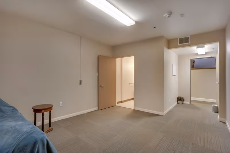 A simple, empty room with beige walls and carpeted floor. There is a small round wooden side table next to a blue-covered piece of furniture on the left. The room has two open doorways leading to other areas, with one door partially open. The ceiling has fluorescent lighting and a ventilation grille above one doorway.