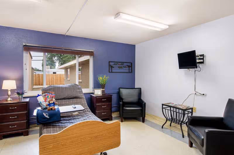 A well-lit patient room with a hospital bed positioned near a large window showing an outdoor view. The room has two dark wooden nightstands with a lamp and decorative items, a black armchair near the bed, another black chair across from it, a small table with magazines, and a wall-mounted TV. The walls are painted in light purple and white tones.