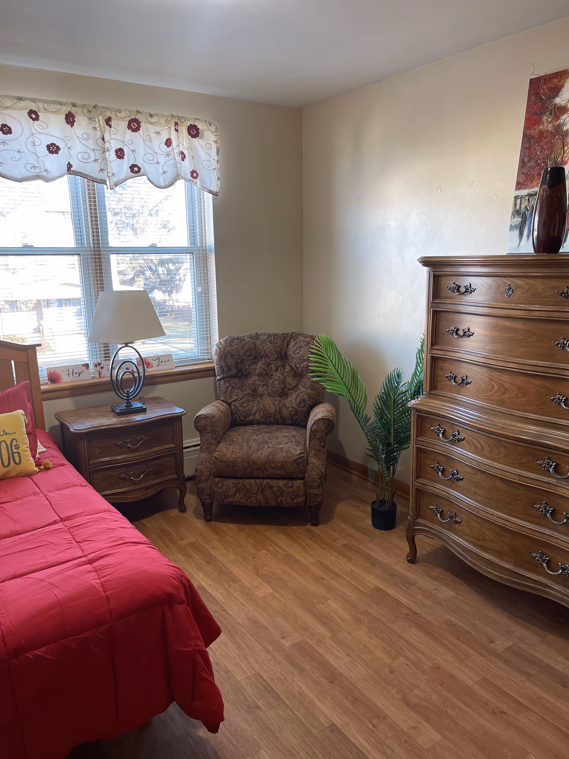 A cozy bedroom with a red bedspread on the bed, a wooden nightstand with a lamp, a patterned armchair, a tall wooden dresser, a potted plant, and a window with floral valance curtains letting in natural light.