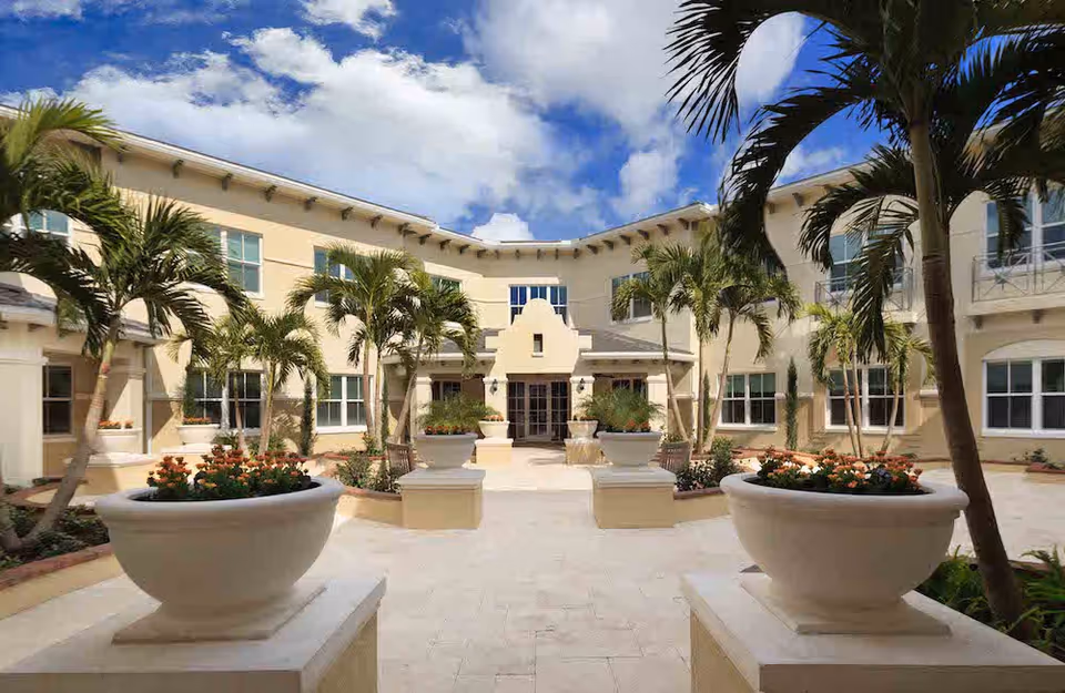 Outdoor courtyard area of a senior living facility with large potted plants, palm trees, and a beige two-story building surrounding the courtyard under a partly cloudy blue sky.