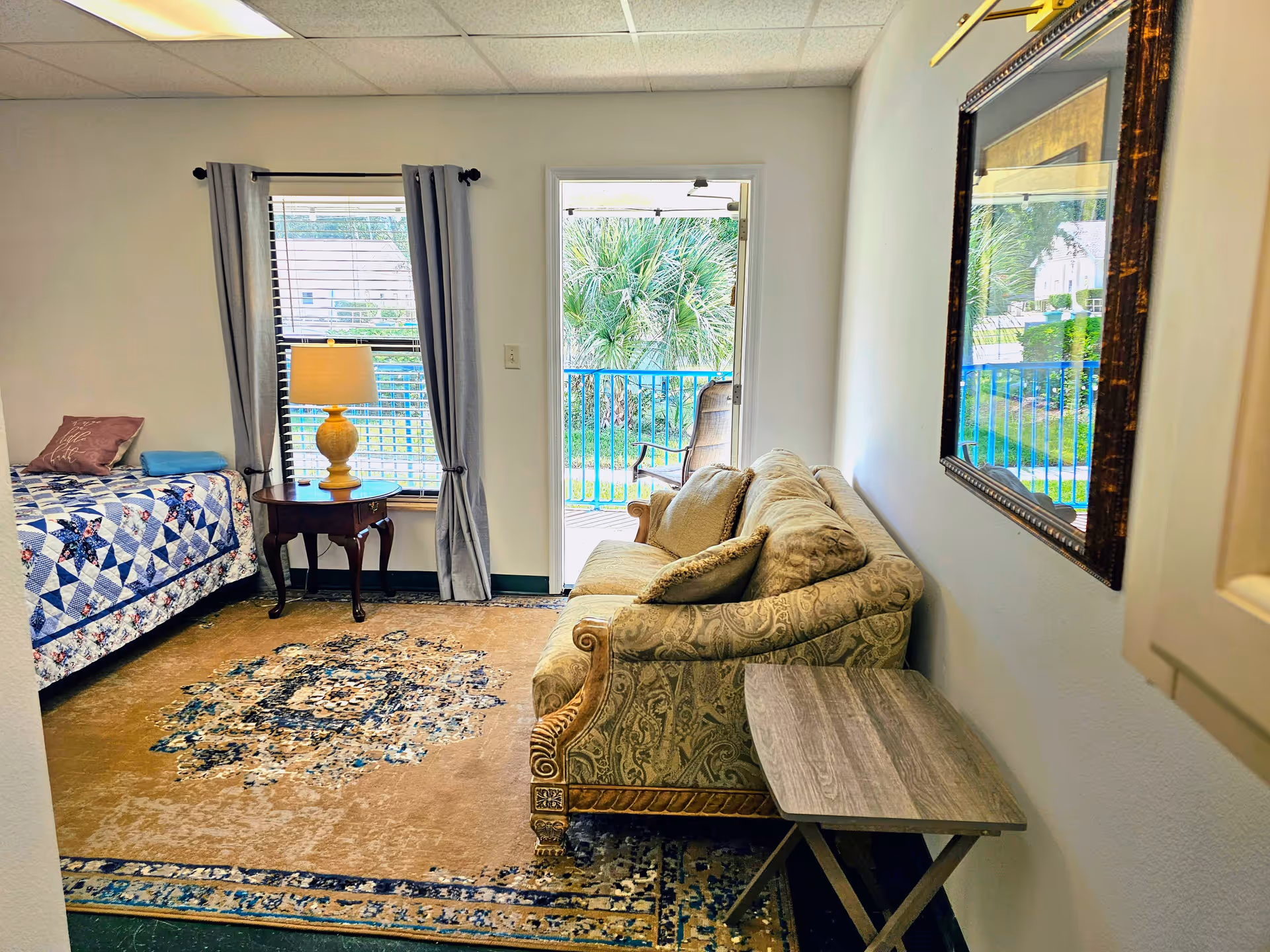 A cozy room with a single bed covered with a blue and white quilt, a wooden side table with a lamp, a patterned beige sofa with cushions, a small wooden folding table, a large decorative mirror on the wall, and a door leading to an outdoor balcony with greenery visible outside.
