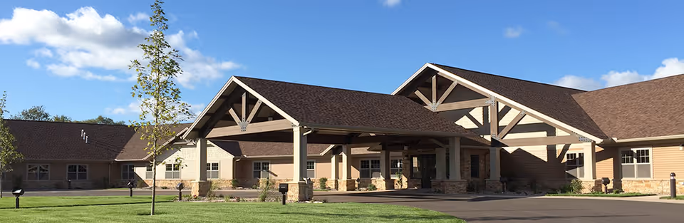 Exterior view of a single-story senior living community building with a large covered entrance, beige siding, stone accents, and a brown roof under a blue sky with scattered clouds. There is a well-maintained lawn and a few young trees in front.