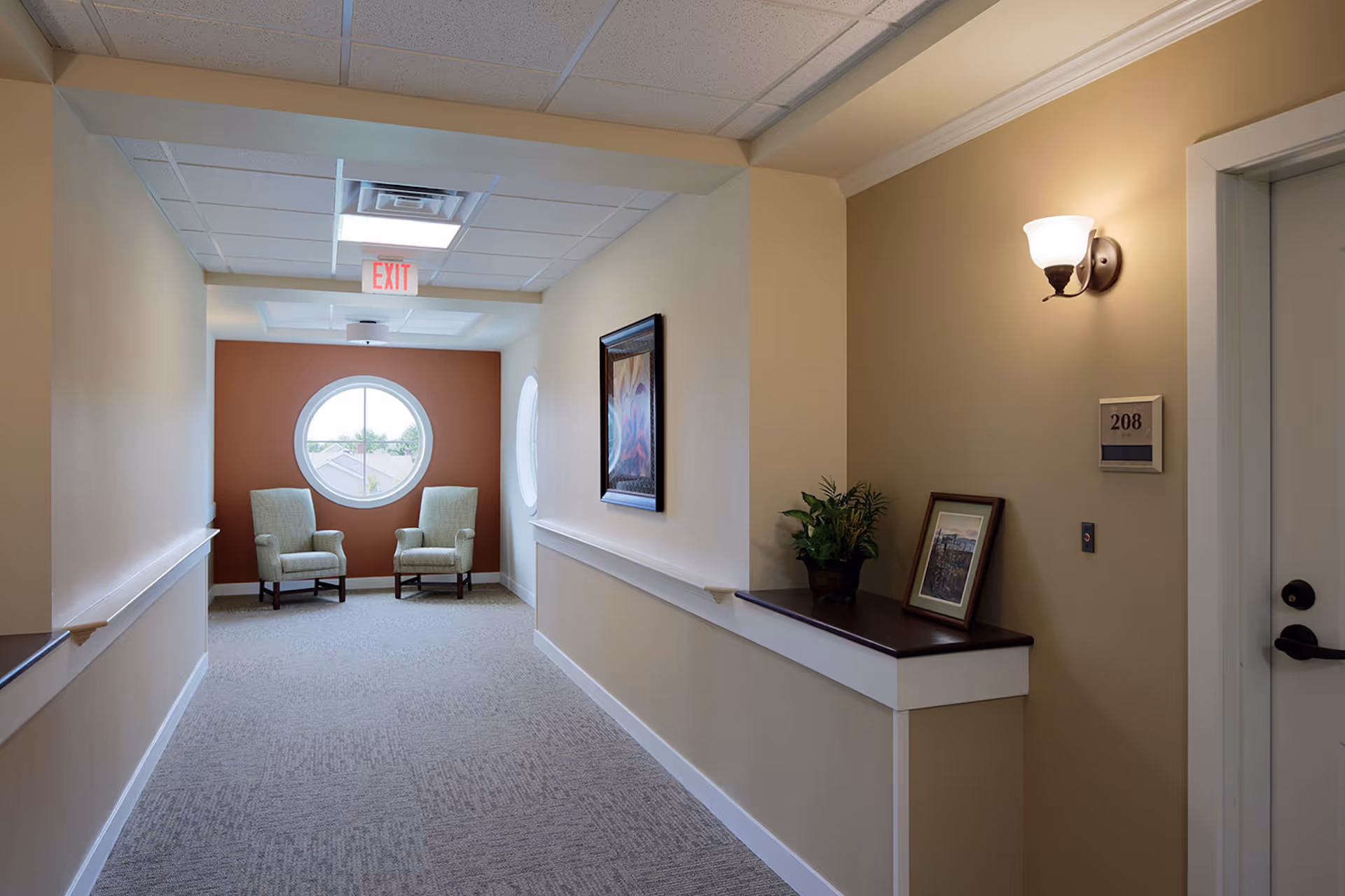 Well-lit carpeted hallway in a senior living facility with two armchairs by a round window, framed art, a console table with a plant, and a door marked 208.