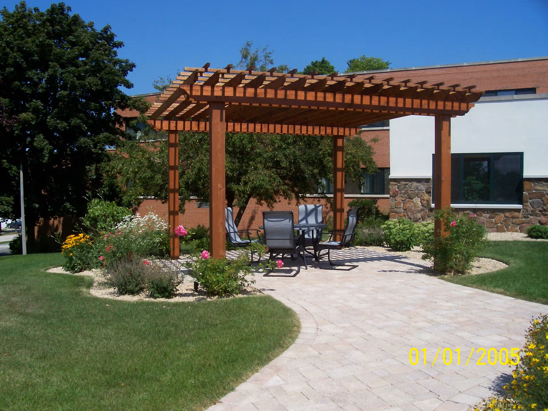 Outdoor patio area with a wooden pergola providing partial shade over a table and four chairs. The area is surrounded by green grass, flower beds with colorful flowers, and trees. A brick and stone building is visible in the background under a clear blue sky.