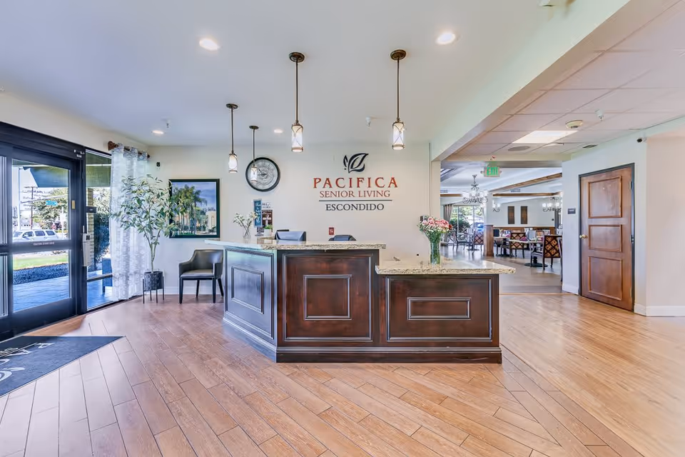 Reception area of Pacifica Senior Living Escondido with a wooden front desk, three pendant lights hanging from the ceiling, a clock on the wall, a plant near the glass entrance door, and a seating area with chairs. The floor is wooden and there is a dining area visible in the background.