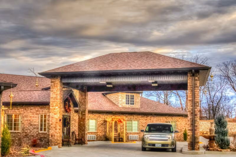 Front exterior of a brick senior living building with a covered porte-cochère and a parked SUV at the entrance.