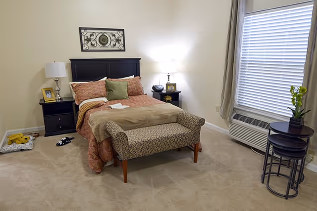 A cozy bedroom with a double bed featuring a dark wooden headboard, red and green pillows, and a beige blanket. There are two bedside tables with lamps, a small bench at the foot of the bed, a window with blinds and curtains, an air conditioning unit below the window, and a small round table with a plant on it. A pet bed with a yellow toy and a pair of slippers are on the carpeted floor.