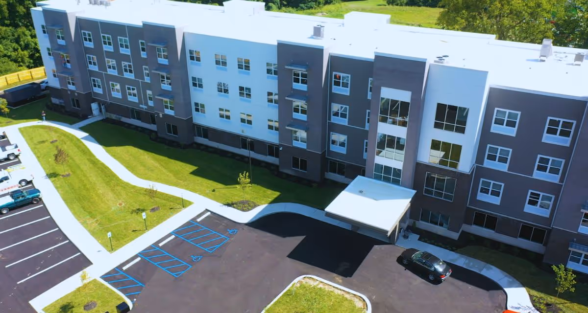 Aerial view of a modern, multi-story senior living facility building with a white and gray exterior. The building is surrounded by green lawns, paved walkways, and a parking lot with several cars parked, including designated handicap parking spaces near the entrance.