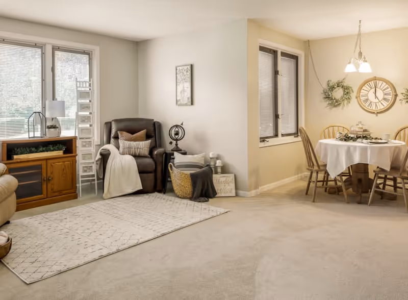 A cozy living and dining area in a senior living facility. The living room features a brown recliner chair with pillows and a throw blanket, a wooden cabinet with a lamp and plant, and a white patterned rug on the carpeted floor. The dining area has a round table covered with a white tablecloth, surrounded by wooden chairs, with a decorative clock and wreaths on the wall.