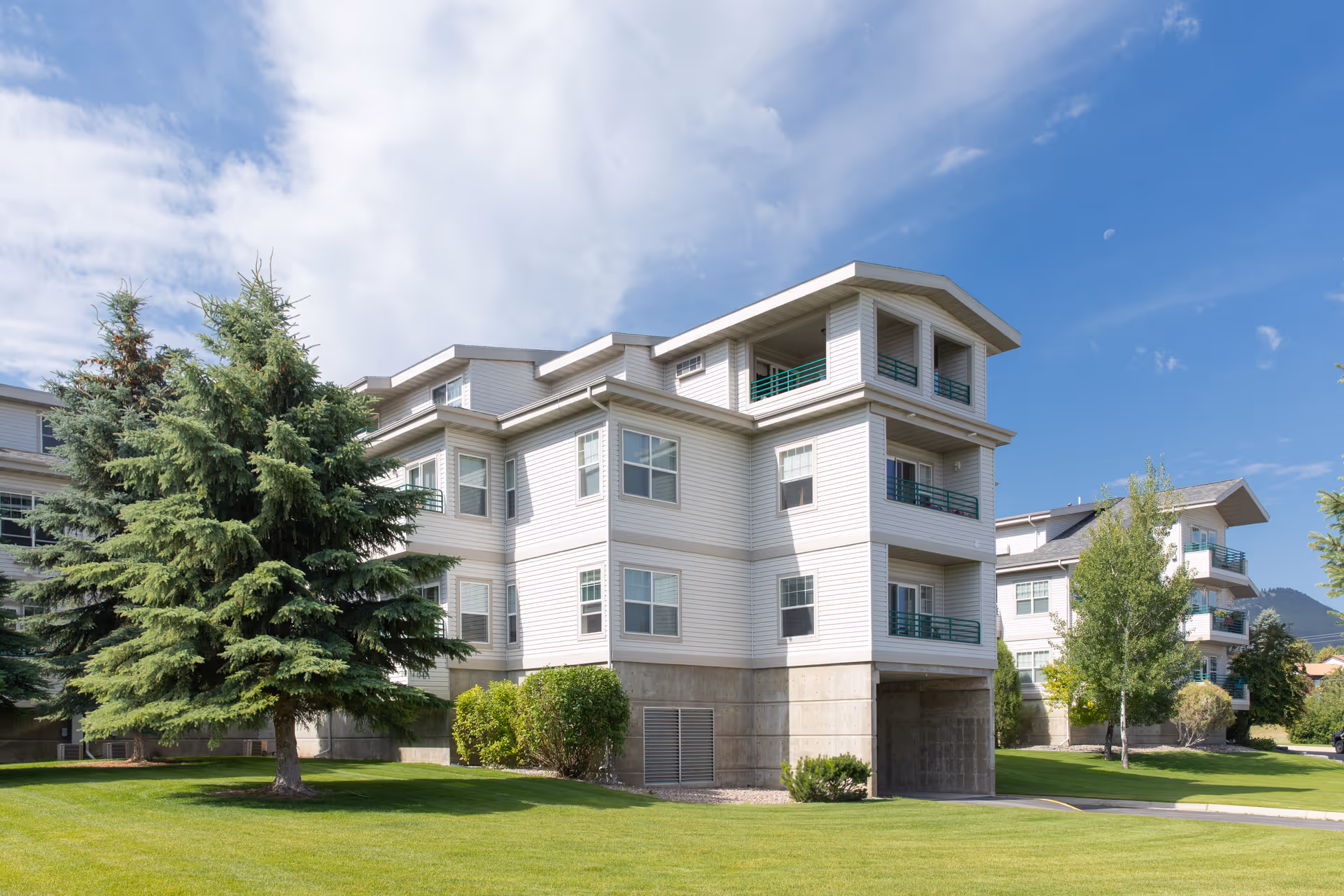 White three-story apartment building with balconies set on a green lawn with trees under a blue sky.