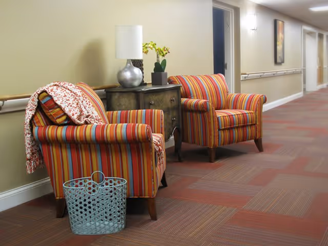 A hallway with two colorful striped armchairs placed against a beige wall. Between the chairs is a small dark wooden table with a silver lamp and a small potted plant. A knitted blanket is draped over one of the chairs, and a blue basket is on the floor in front of it. The hallway has a patterned carpet and handrails along the wall, with several doors and framed artwork visible in the background.