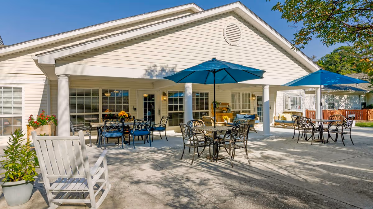 Outdoor patio area at Brookdale Roanoke featuring multiple metal tables with chairs and blue umbrellas providing shade. The patio is adjacent to a light-colored building with large windows and a covered porch supported by white columns. There are potted plants and a white rocking chair on the concrete patio under a clear blue sky.
