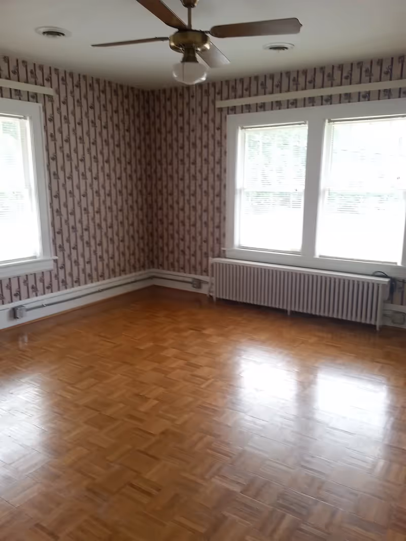Empty room with wooden parquet flooring, floral patterned wallpaper, two large windows with blinds, a radiator below the windows, and a ceiling fan with a light fixture.