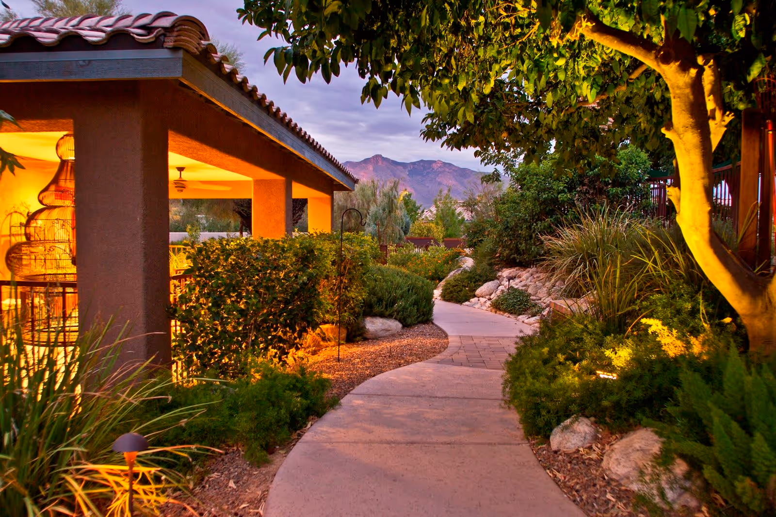 A winding outdoor pathway surrounded by lush greenery, bushes, and trees leading past a building with a tiled roof and warm interior lighting. Mountains are visible in the background under a cloudy sky.