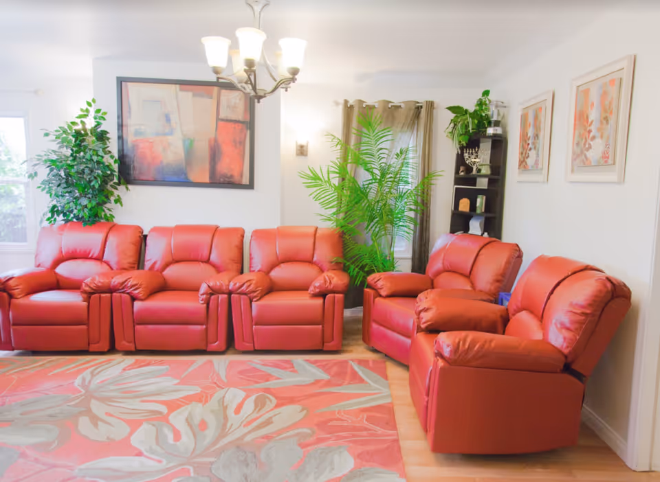 A bright living room with five red leather recliner chairs arranged along the walls. There is a large colorful abstract painting on the wall, two windows with curtains, several green plants, a black shelving unit with decorative items, and a chandelier hanging from the ceiling. The floor has a large area rug with a floral pattern in shades of red, beige, and gray.