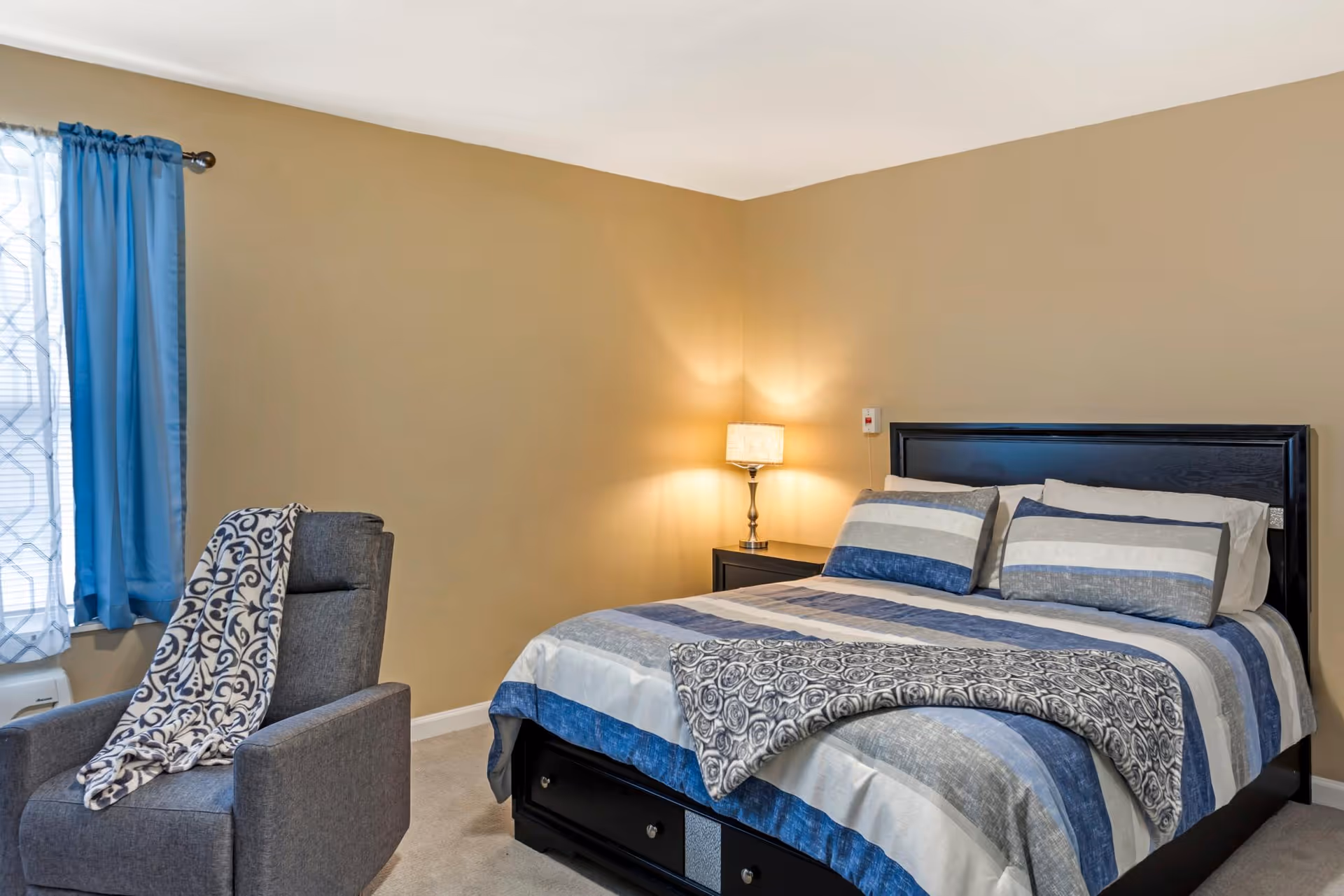 A bedroom with a neatly made bed featuring blue, gray, and white striped bedding. There is a black headboard and a matching black nightstand with a lamp on it. A gray armchair with a patterned blanket draped over the back is positioned near a window with blue curtains.