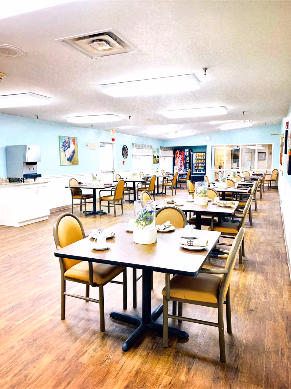 A bright dining room with multiple tables and chairs arranged neatly. Each table has place settings with cups, plates, and napkins. The room has light blue walls, wood flooring, and fluorescent ceiling lights. There is a vending machine and a water dispenser along the walls, and some artwork and decorations are visible on the walls.