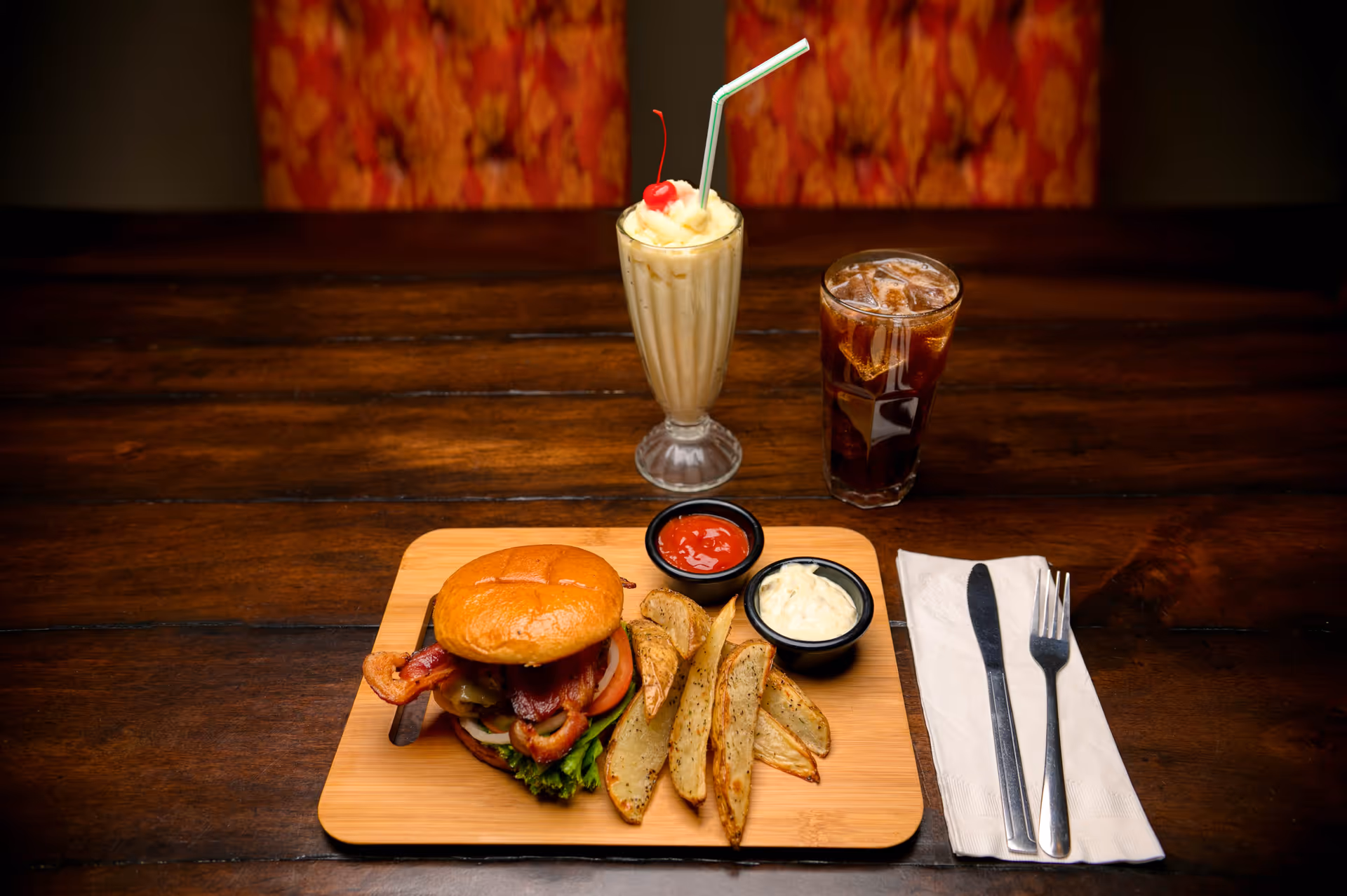 A wooden table with a meal consisting of a bacon cheeseburger, potato wedges with ketchup and mayonnaise dips, a glass of iced cola, and a vanilla milkshake topped with whipped cream and a cherry, with a knife and fork on a napkin beside the meal.