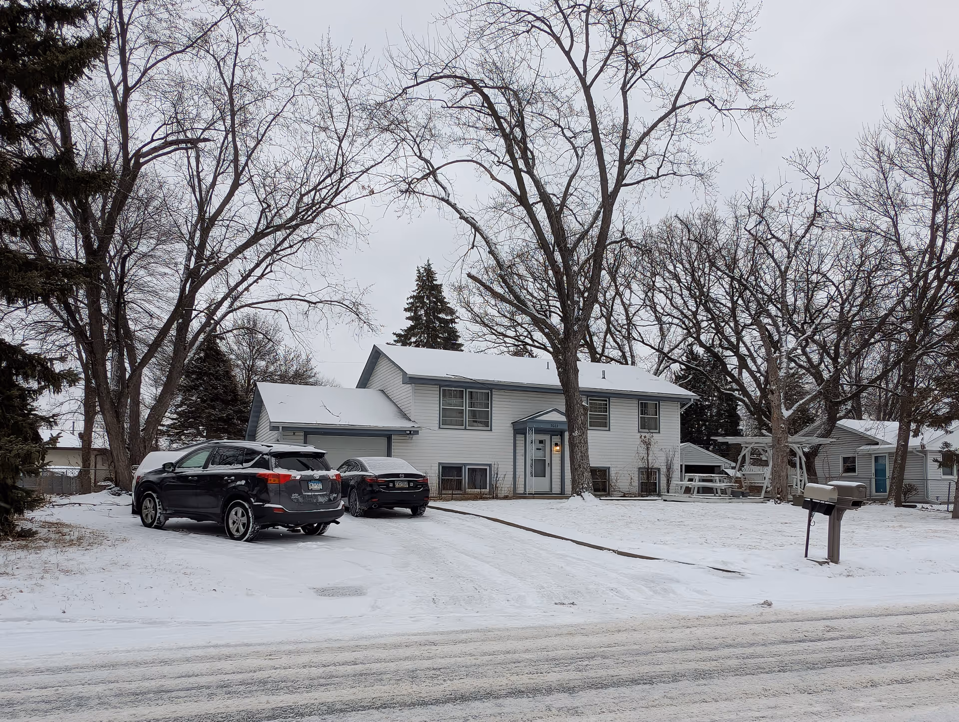 Snow-covered driveway and yard in front of a two-story light-gray house with two parked cars and leafless trees.