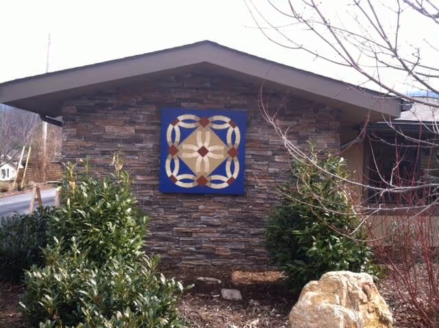 Exterior view of a building with a stone facade featuring a decorative square quilt-like pattern mounted on the wall. There are green bushes and a large rock in the foreground, with leafless tree branches visible on the right side.