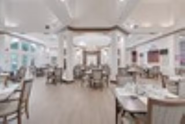 A spacious dining room with multiple tables and chairs arranged neatly. The room features large windows allowing natural light to fill the space, light-colored walls, and a wooden floor. The ceiling has recessed lighting and decorative molding, creating a welcoming atmosphere.