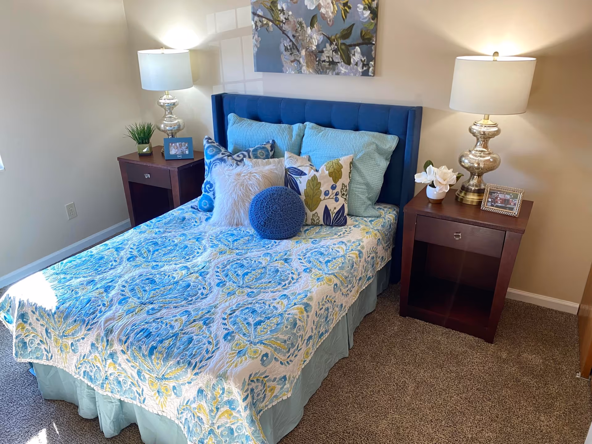 Small bedroom with a neatly made bed featuring blue patterned bedding, multiple decorative pillows, a tufted blue headboard, and two bedside tables with lamps.