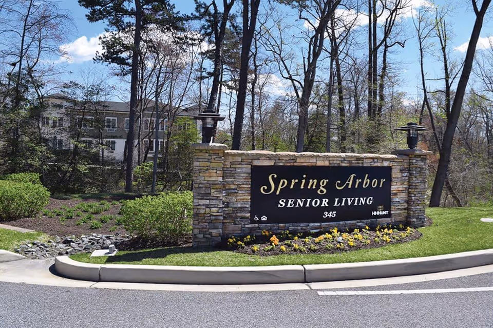 Stone sign at the entrance of Spring Arbor Senior Living with the address number 345, surrounded by landscaped greenery and flowers, with trees and a building visible in the background under a blue sky.