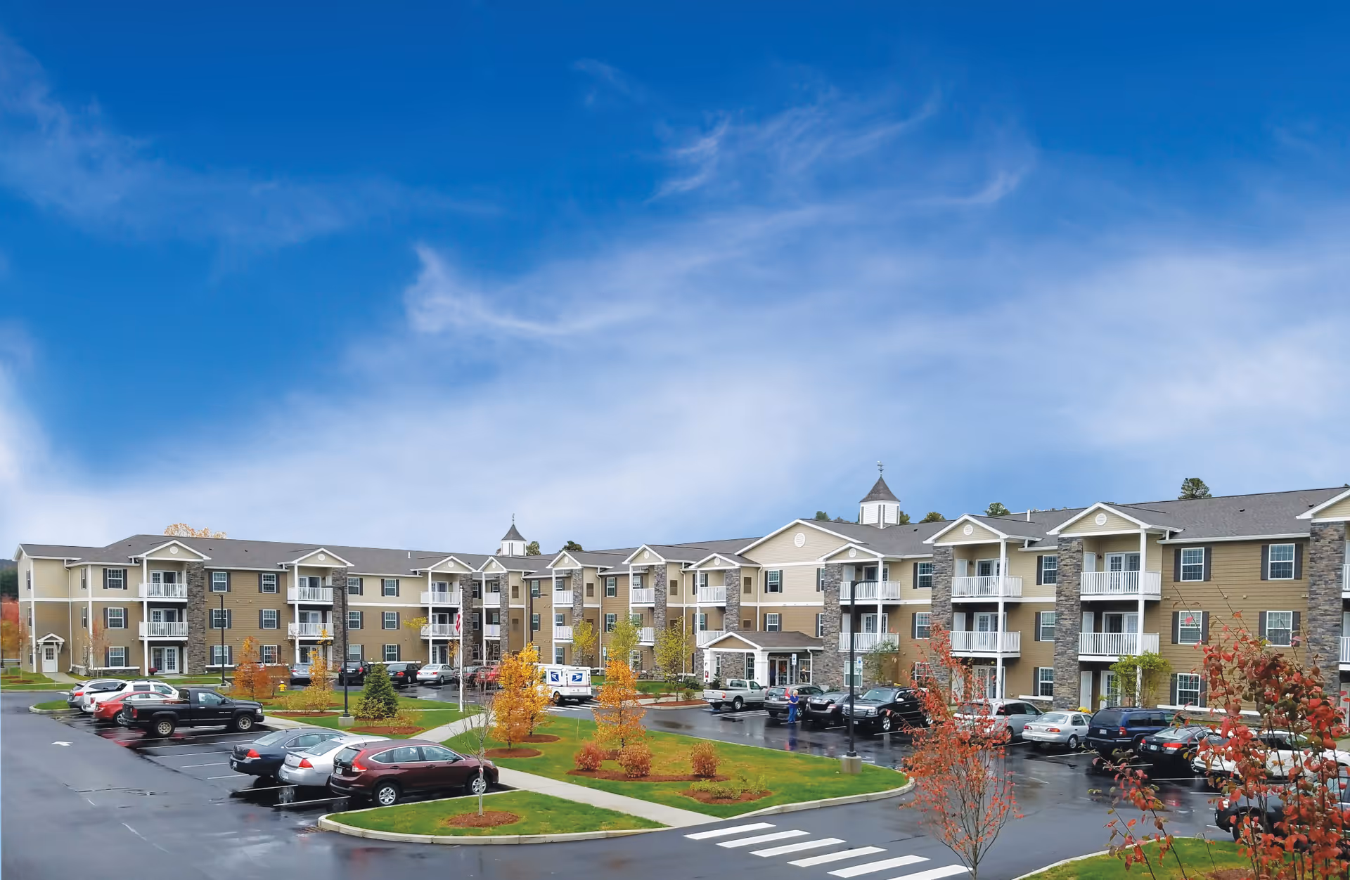 Exterior view of a three-story active adult retirement community building with balconies, surrounded by a parking lot with cars and landscaped areas with small trees and grass under a partly cloudy blue sky.