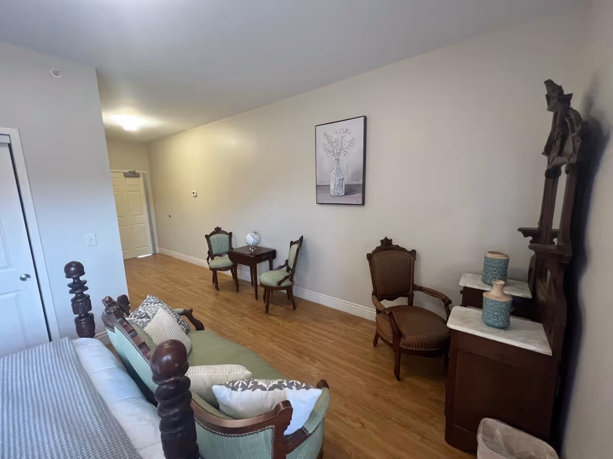 A room with wooden flooring featuring a vintage-style wooden bed with green upholstery and pillows, two antique chairs with green upholstery, a small wooden table with a globe on it, a wooden dresser with a mirror and decorative vases, and a framed picture of a vase with flowers on the wall.