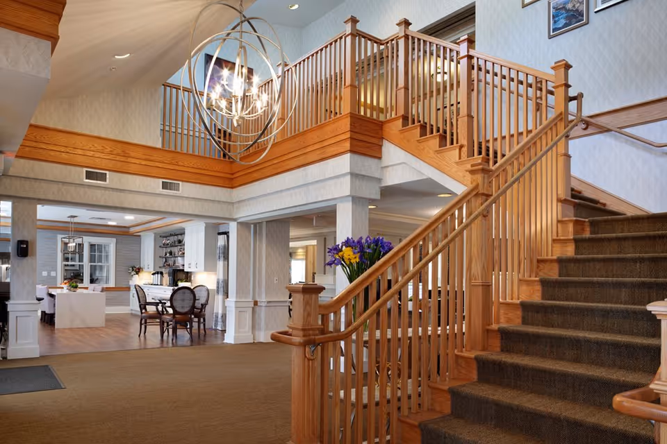 Spacious interior lobby featuring a carpeted wooden staircase, large chandelier, and an open dining/kitchen area.