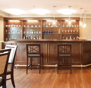 Interior view of a wooden bar area with two high chairs in front. Behind the bar are shelves displaying various glassware and decorative items. The floor is wooden, and there are pendant lights hanging from the ceiling above the bar.